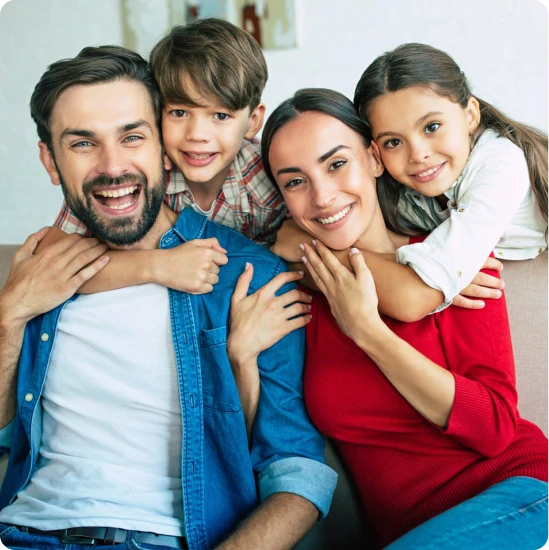 A happy family of four, including a man, woman, young boy, and young girl, sitting closely together on a couch in Katy Tx, smiling and posing for the camera—protected with Casillas Insurance Services.