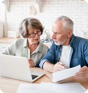 An older couple sits at a table with a laptop and paperwork, looking concerned as they review documents together in their bright Katy, TX kitchen, possibly considering Insurance Services from Casillas Insurance.
