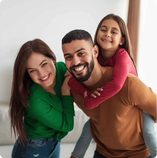 A smiling man with a beard carries a young girl on his back while a woman stands beside them. They all look happy and are posing closely together indoors, capturing a joyful family moment in Katy TX, home to Casillas Insurance services.