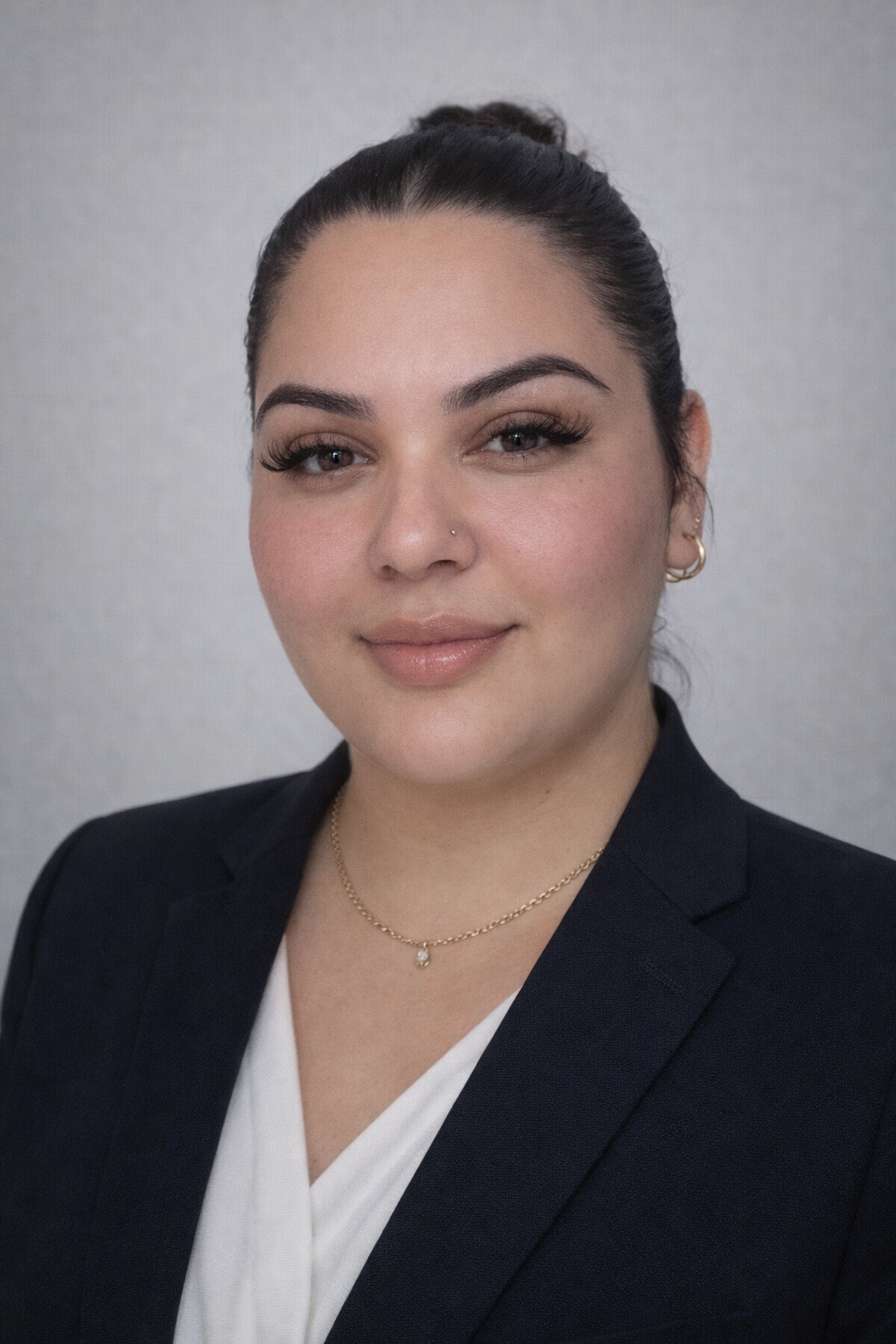A woman with dark hair in a bun, wearing a dark blazer, white top, gold necklace, and hoop earrings, smiles slightly in front of a plain light gray background—representing Casillas Insurance in Katy, TX.