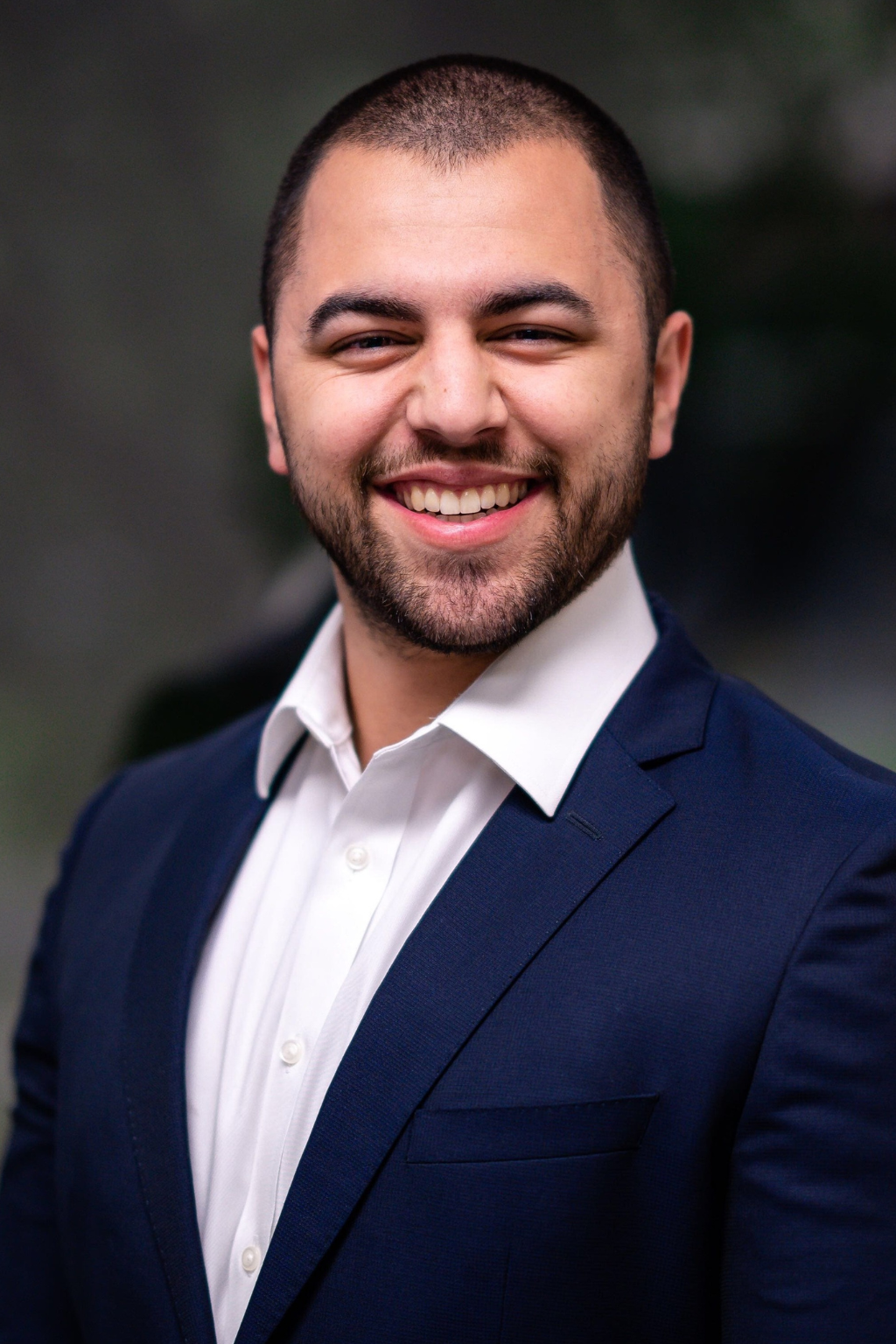 A man in a navy blue suit jacket and white shirt smiles confidently at the camera against a blurred outdoor background, representing Casillas Insurance and their trusted insurance services in Katy, TX.