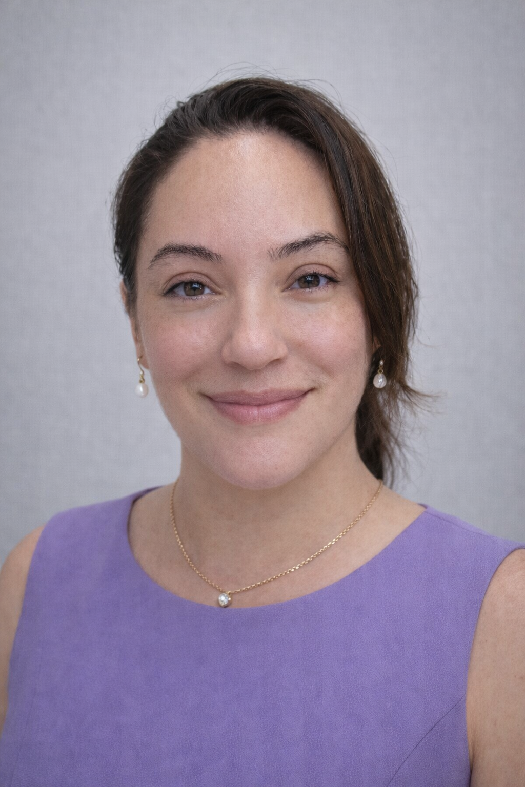 A woman with light skin and straight brown hair smiles gently. She is wearing a sleeveless lavender top, pearl earrings, and a delicate gold necklace. The background is plain light gray, reflecting the warm professionalism of Casillas Insurance in Katy TX.