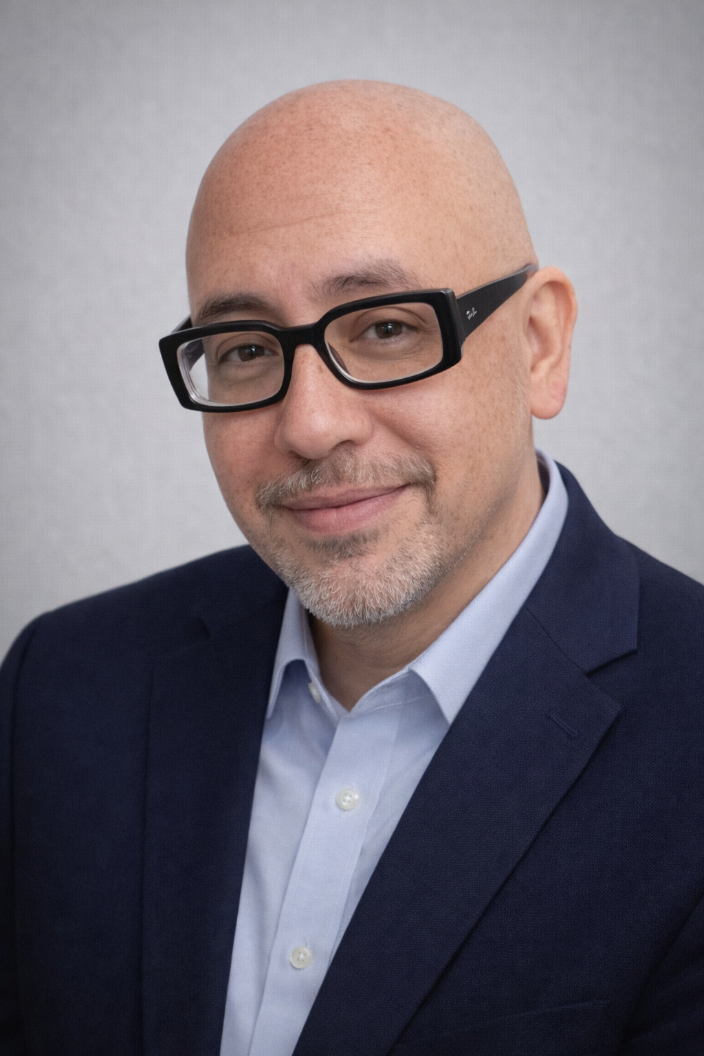 A bald man with glasses, a short beard, and a slight smile wears a navy blue suit jacket and light blue shirt, standing in front of a plain light gray background—representing Casillas Insurance services in Katy Tx.